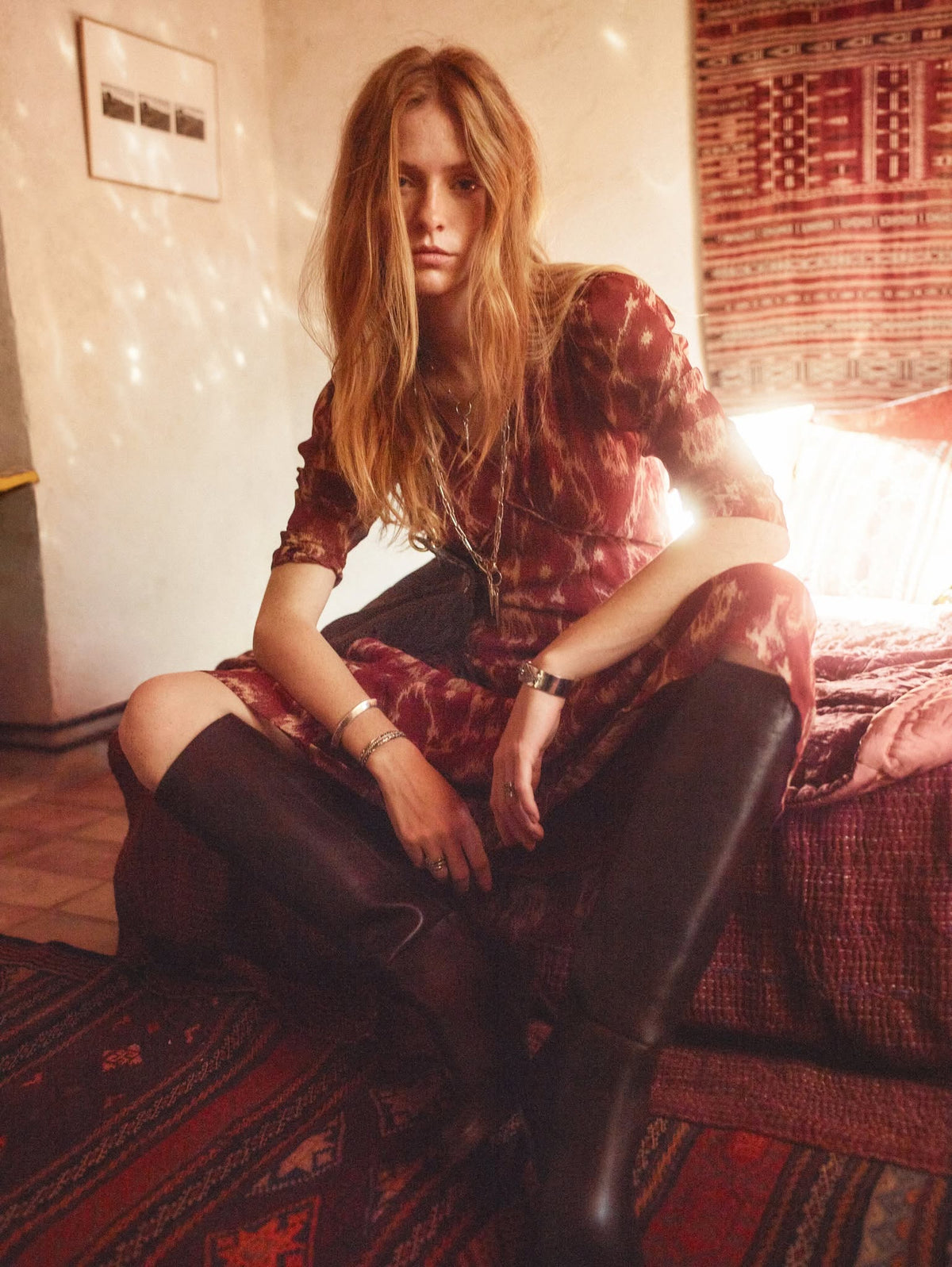 Woman in printed Ba&sh dress and tall boots seated on patterned bedspread
