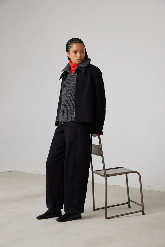 Model in black jacket and wide-leg trousers over grey knit, hand on metal chair
