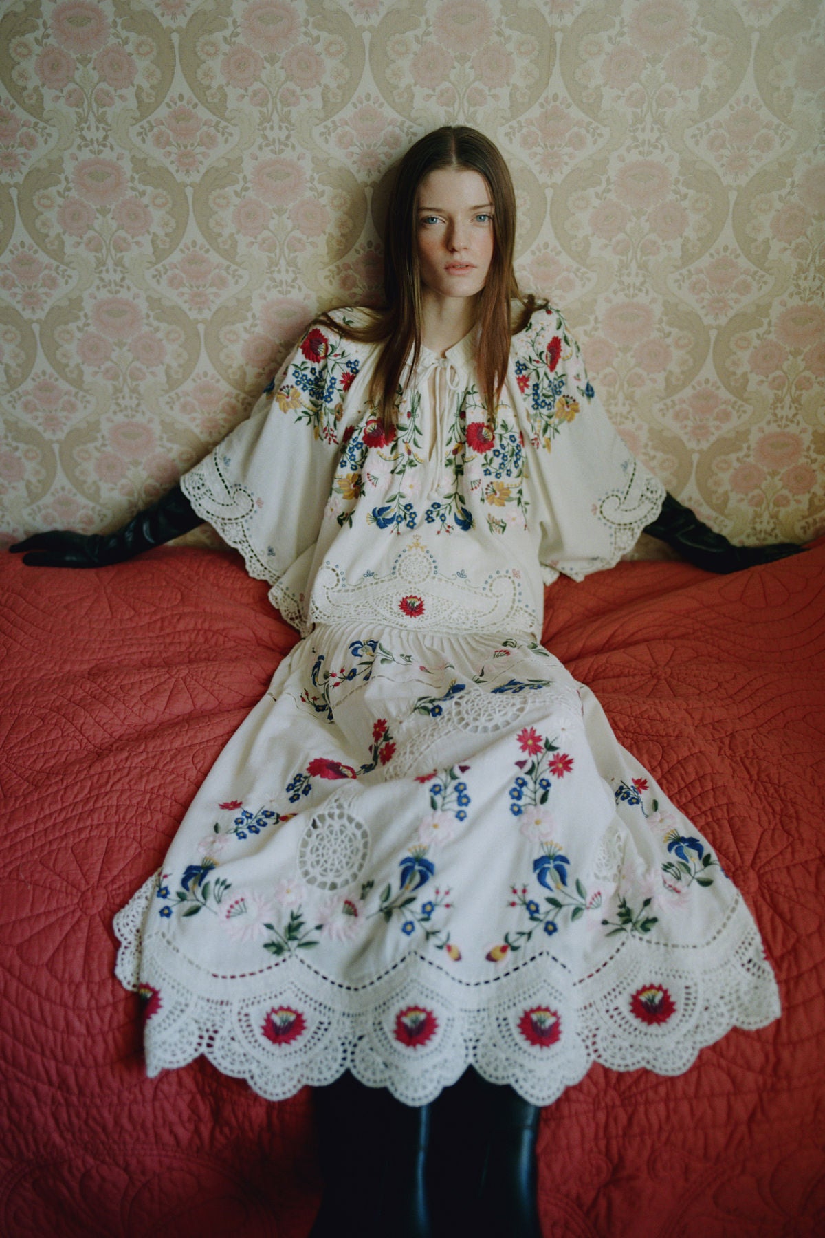 Model in floral embroidered dress with crochet lace hem, seated on red bedspread