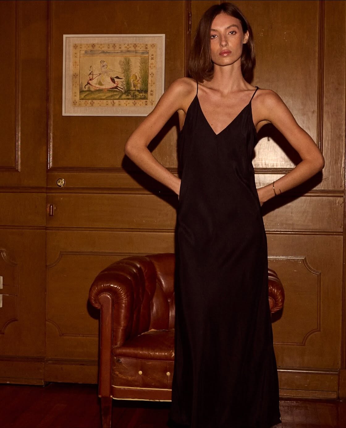 Model wears a black spaghetti-strap maxi slip dress in a wood-panelled room