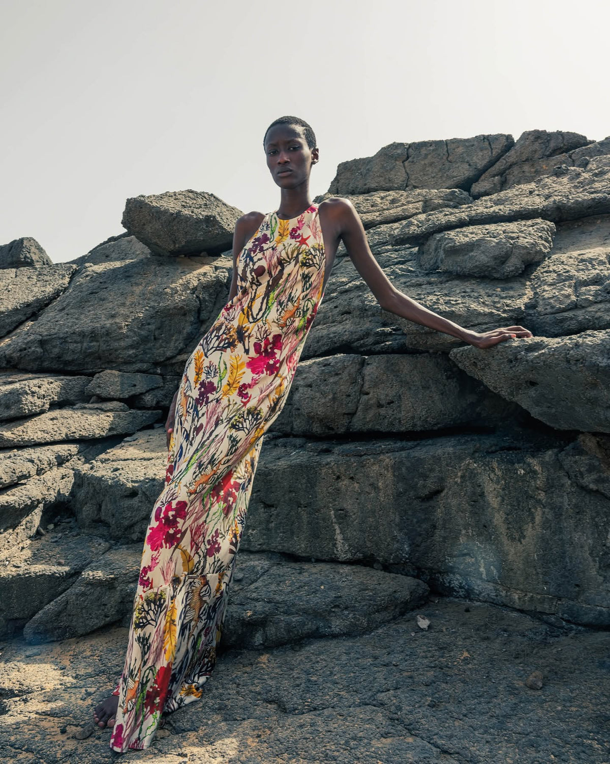 Model wears a floral-print silk maxi dress, leaning against large coastal rocks