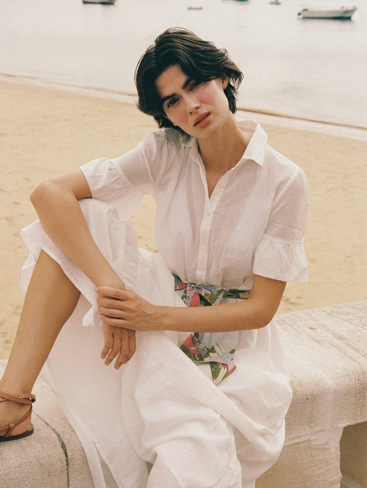 Woman in white shirtdress with floral sash sits on a seaside wall