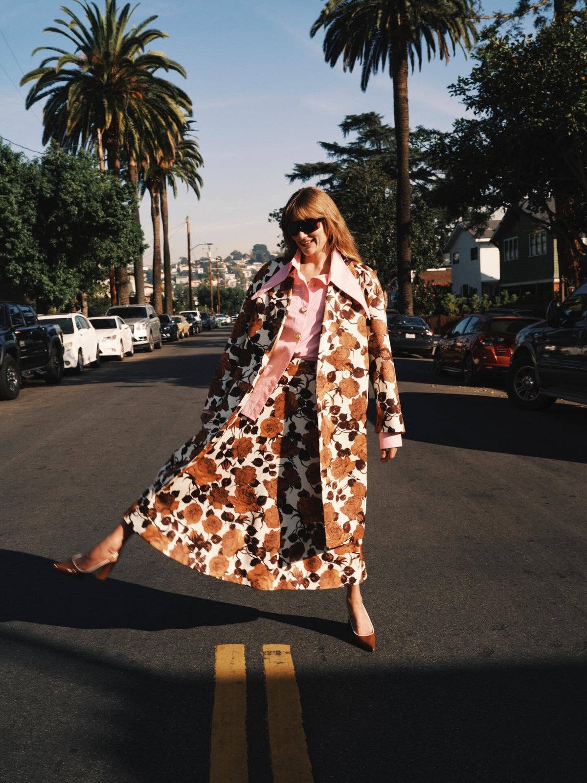 Woman in brown floral maxi coat and pink shirt walking a palm-lined street