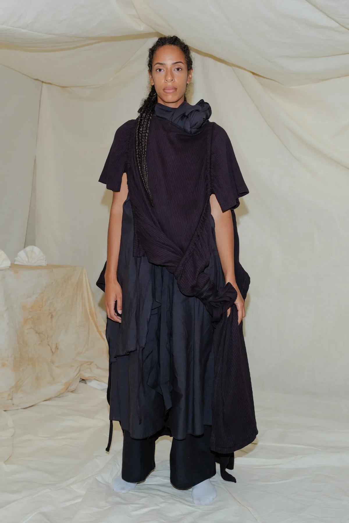 Model wears a draped dark top and layered black skirt with twisted scarf detail.