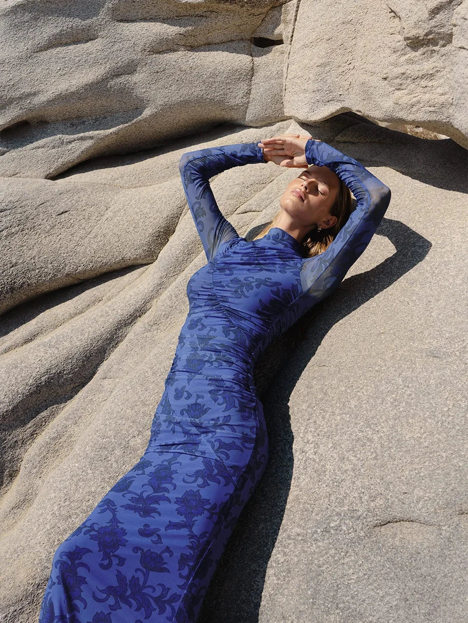 Woman in long-sleeve blue floral maxi dress reclining on sunlit rocks