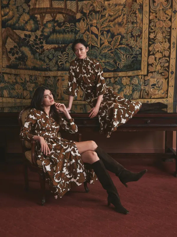 Two models in matching brown paisley-print dresses before a tapestry backdrop