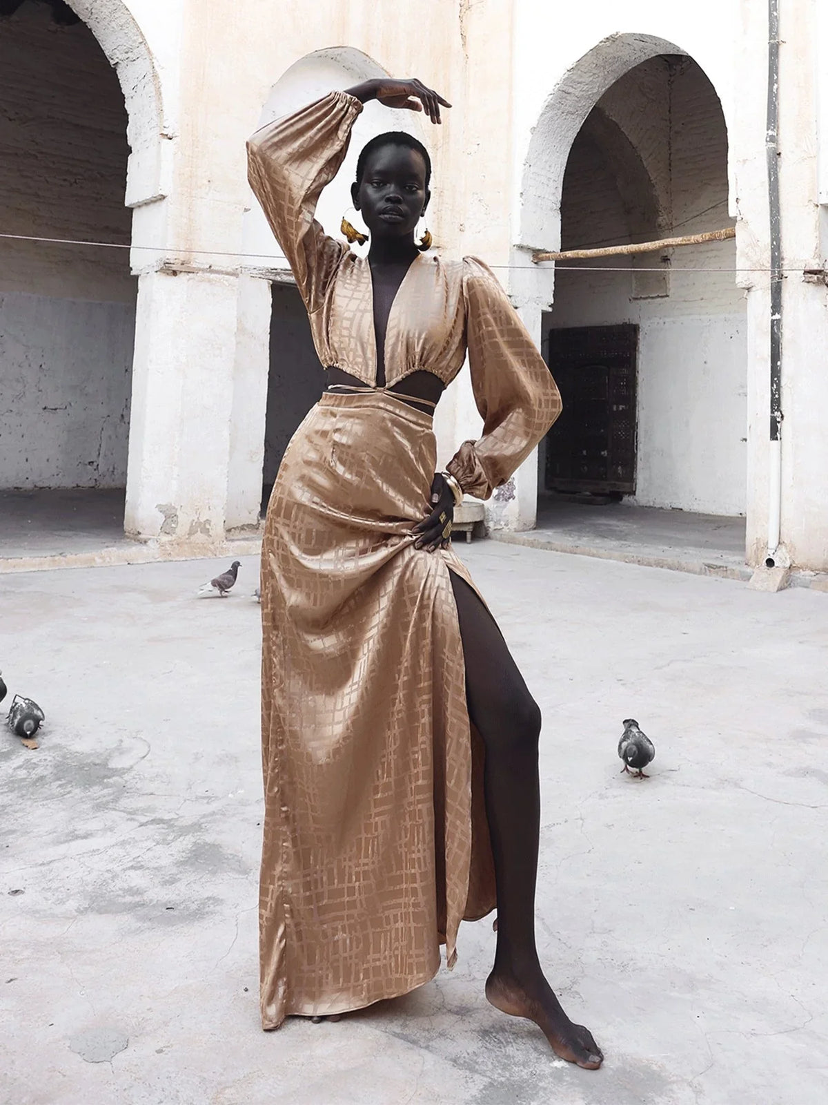 Model wears a plunging gold satin gown with cutout waist and thigh slit, arm raised