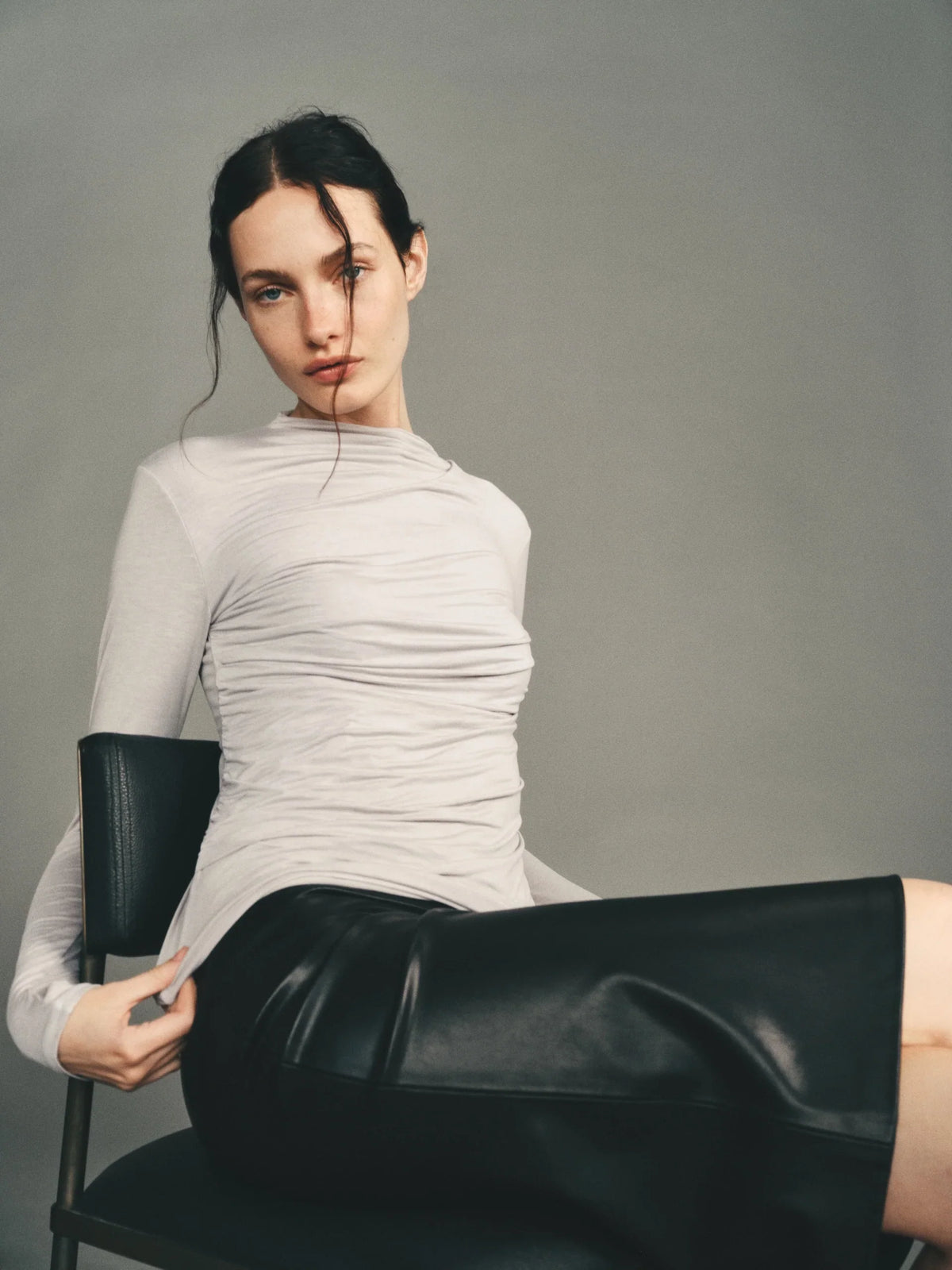 Model in ruched long-sleeve top and black leather skirt, seated on a chair