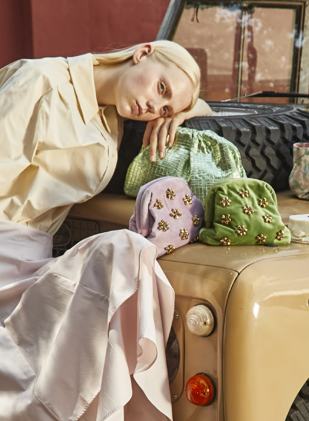 Model resting on a vintage car beside green and lilac embellished clutch bags