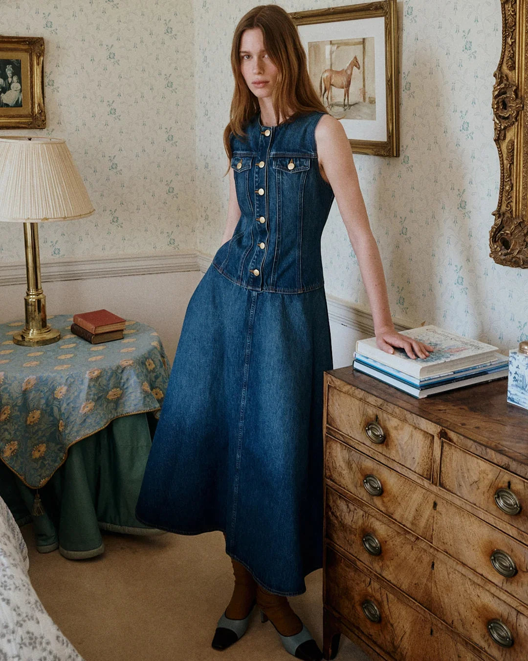 Woman in sleeveless denim bodice and full midi skirt beside antique wooden dresser