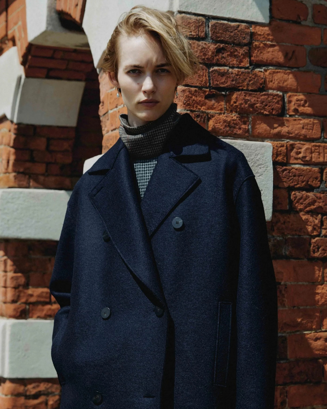 Model in oversized navy double-breasted coat over a check turtleneck, against a brick wall
