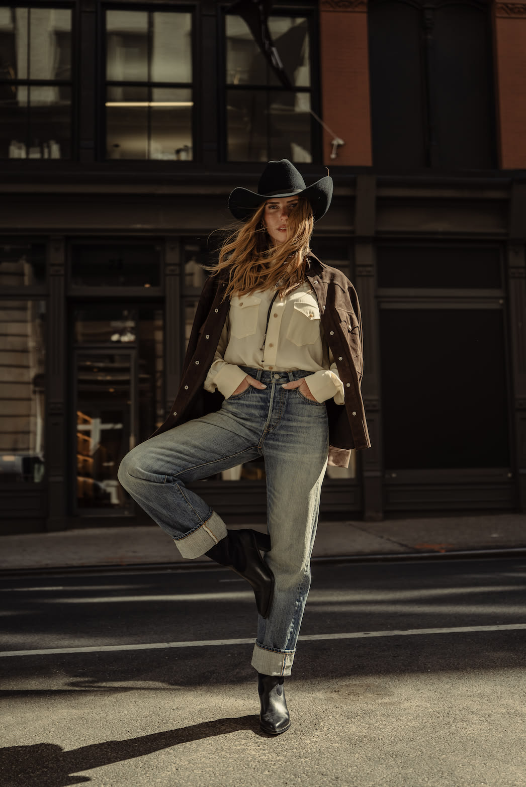 Woman in cowboy hat, cuffed jeans and black ankle boots posing on city street
