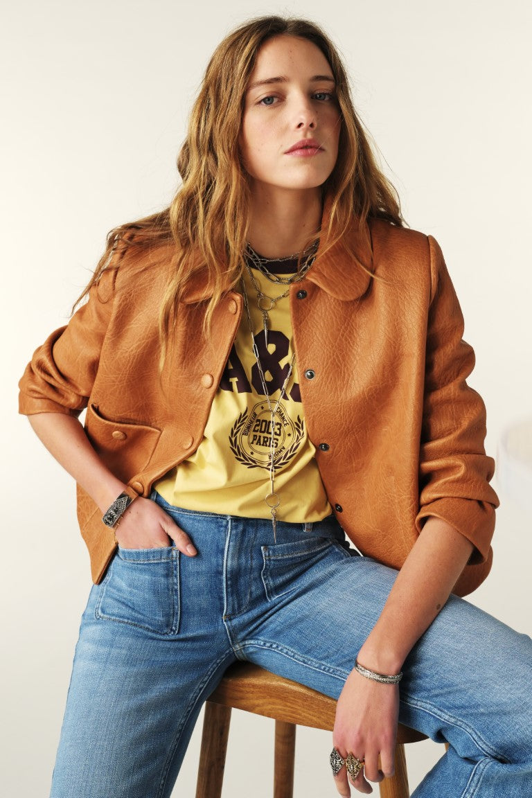 Woman wearing a brown jacket, yellow graphic t-shirt, and blue jeans sitting on a wooden stool.