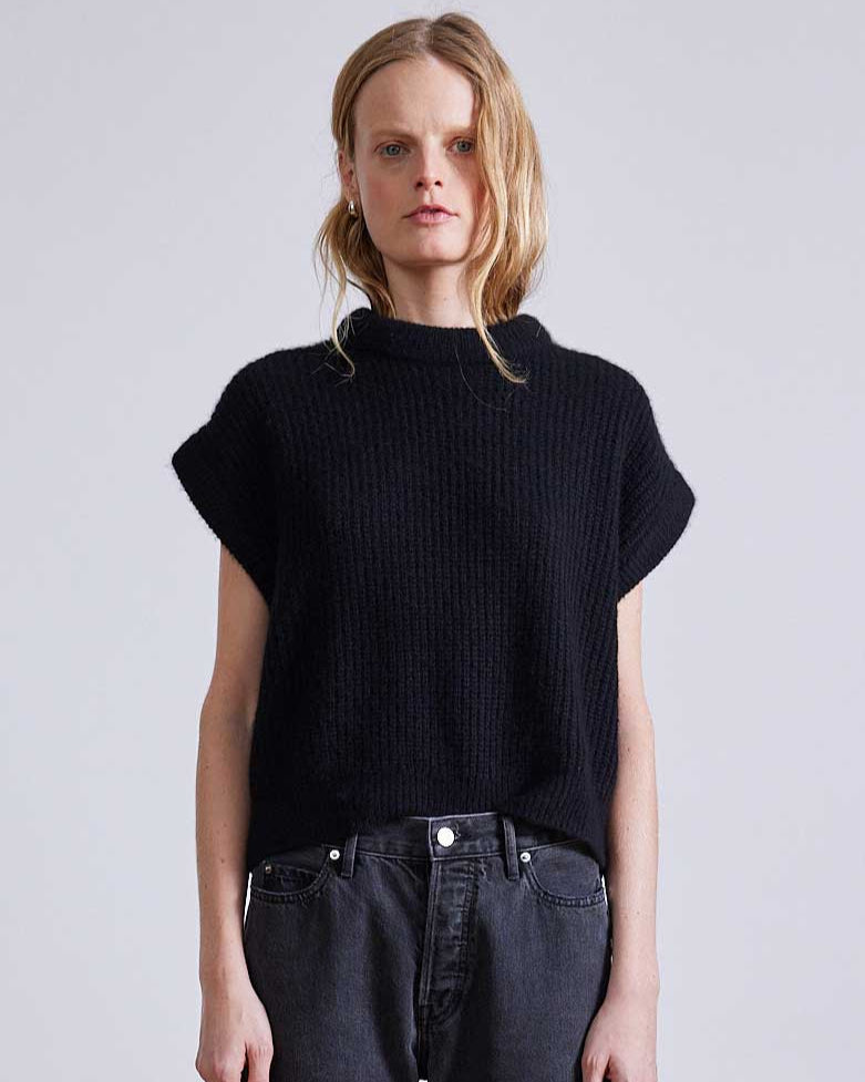 Woman wearing a black knit top and dark jeans against a plain background