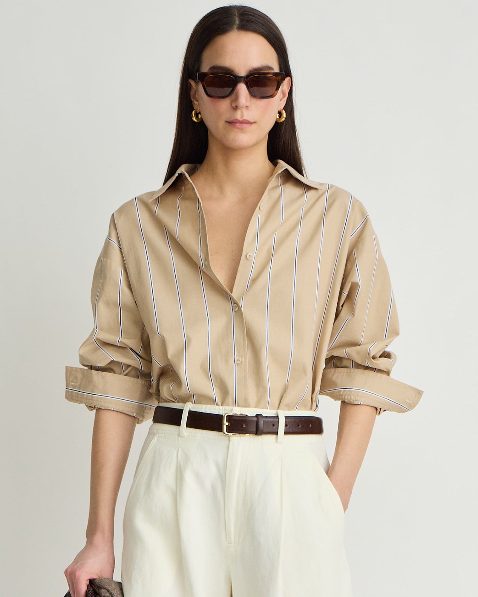 Woman wearing a beige striped shirt and white pants with sunglasses on a plain background