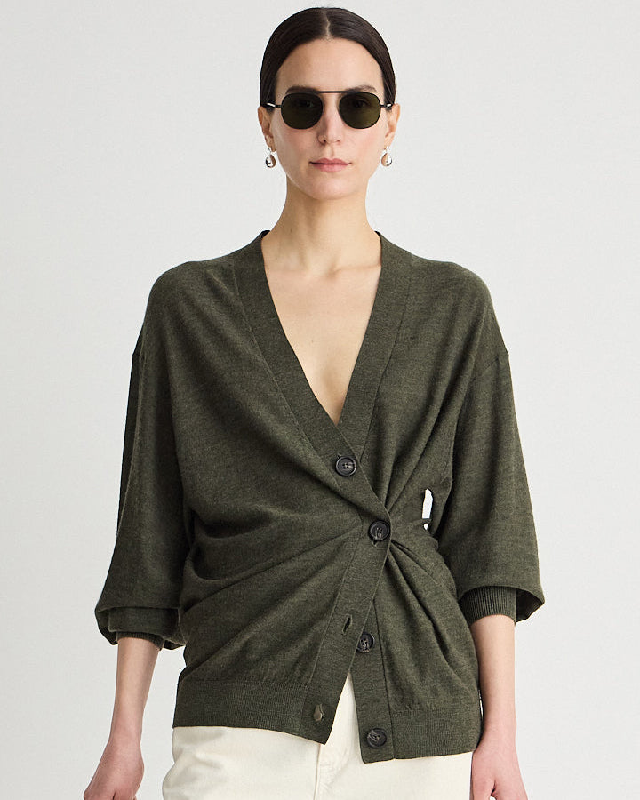 A woman modeling an olive green knit wrap cardigan with a twisted front detail and dark buttons, paired with cream pants and sunglasses, against a light gray studio background.