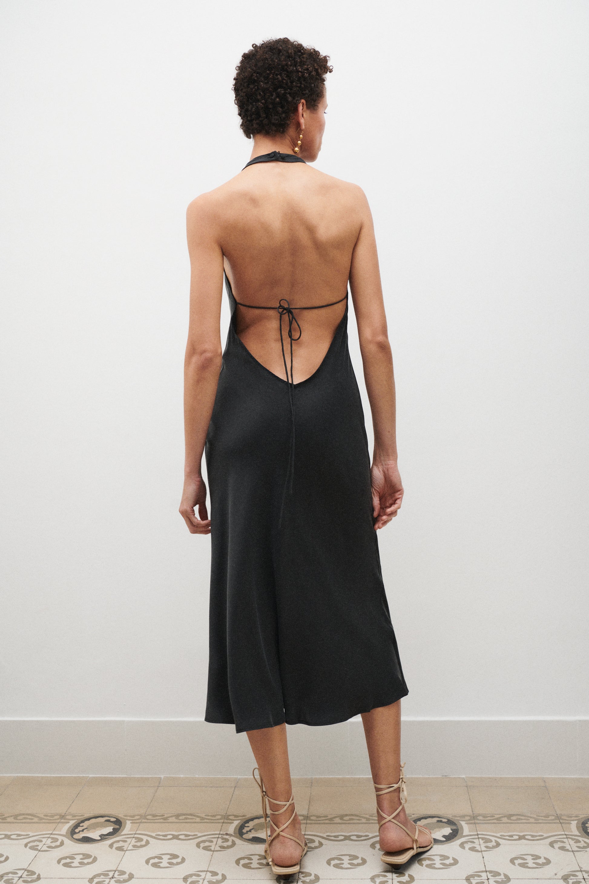 Woman wearing a black dress with an open back, standing against a white wall.