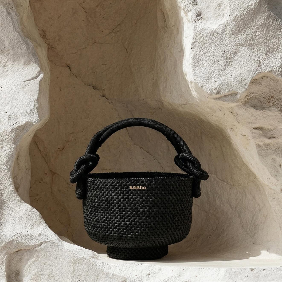 A black woven bucket handbag with a knotted rope handle, displayed on a white stone shelf against a textured white rock wall.