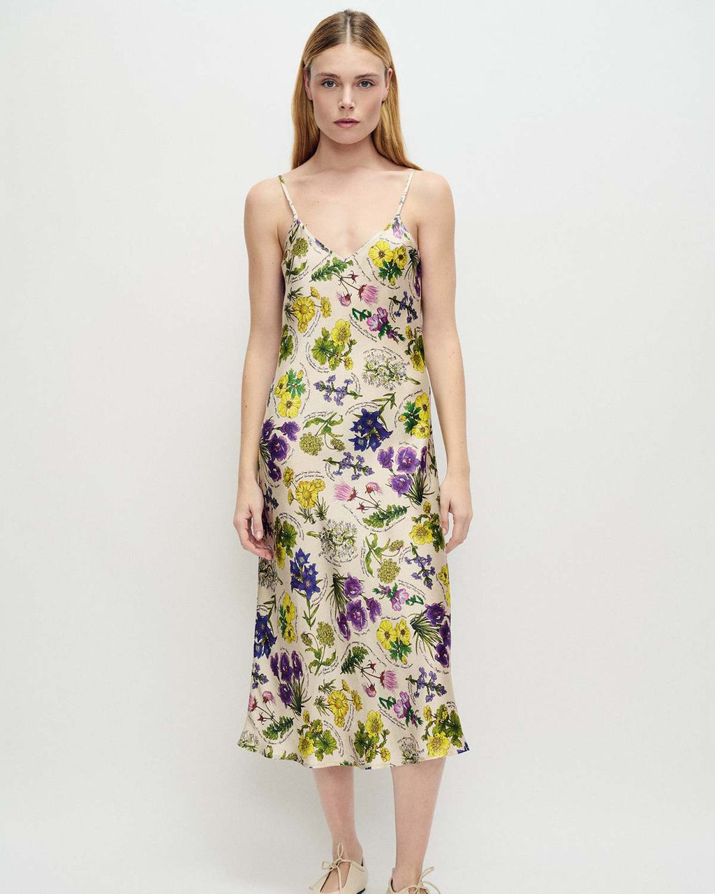 A model stands against a plain white studio background wearing a sleeveless midi slip dress with thin straps and an all-over floral print in purple, yellow, green, and blue on a light base. She is also wearing beige flat shoes.