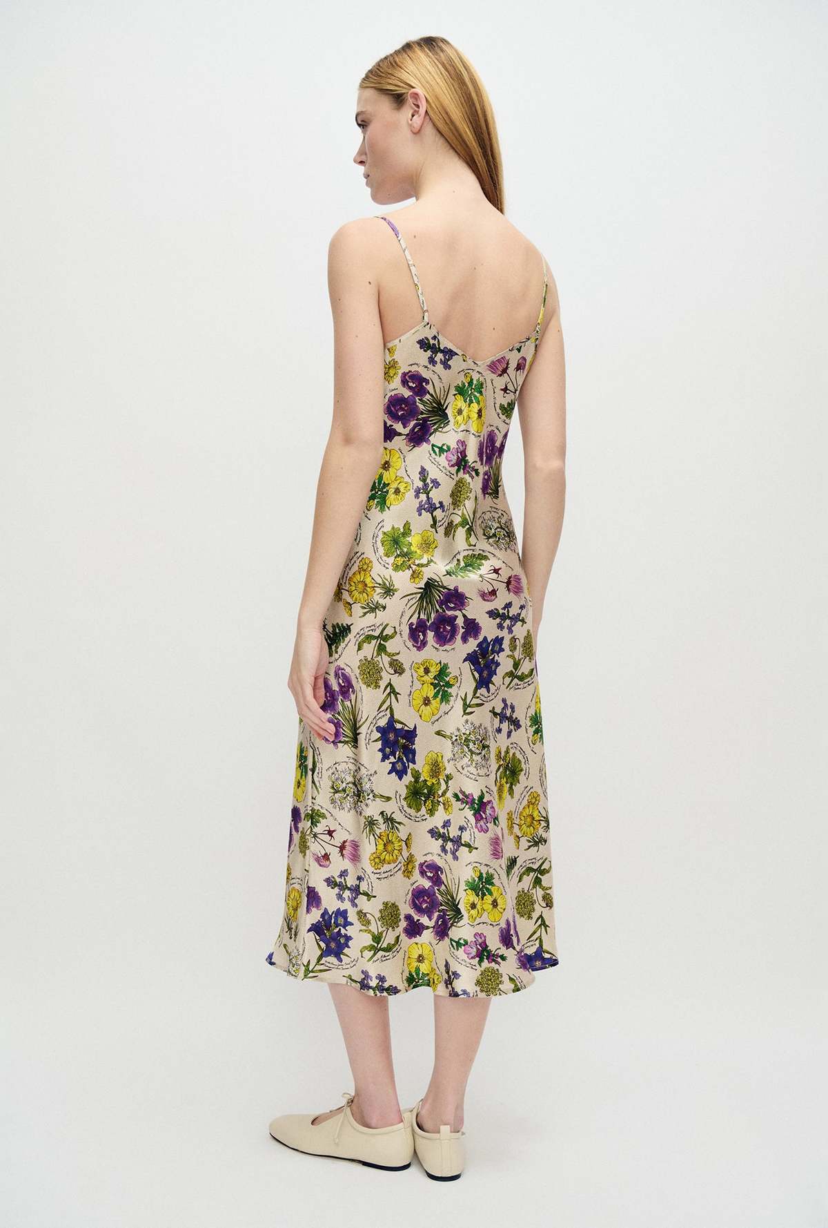 A woman shown from the back wearing a sleeveless midi dress with thin straps and a colorful floral print on a light beige base. She is standing against a plain white studio background.