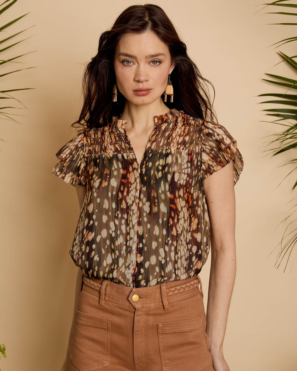 Woman wearing a patterned blouse and brown pants against a beige background with green leaves.