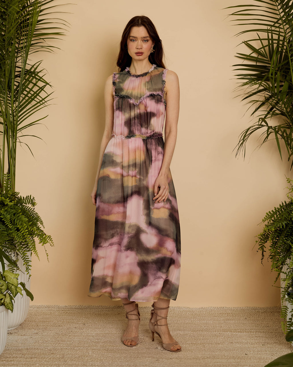 A woman stands in a studio setting wearing a sleeveless midi dress with an abstract watercolor-like print in pink, beige, gray, and olive tones. The background is a warm beige wall with potted palm plants framing the scene.