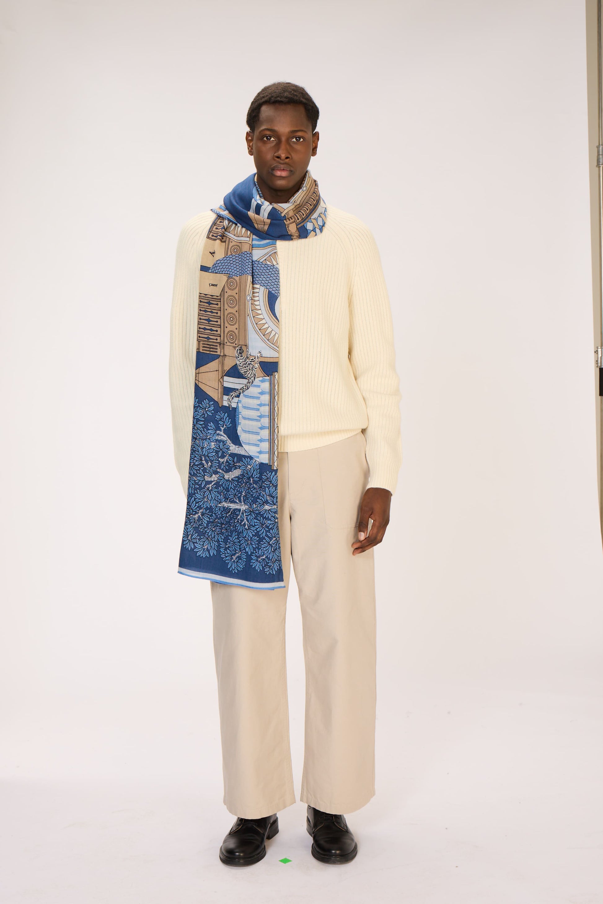 Man wearing a cream sweater, patterned scarf, and beige pants on a white background