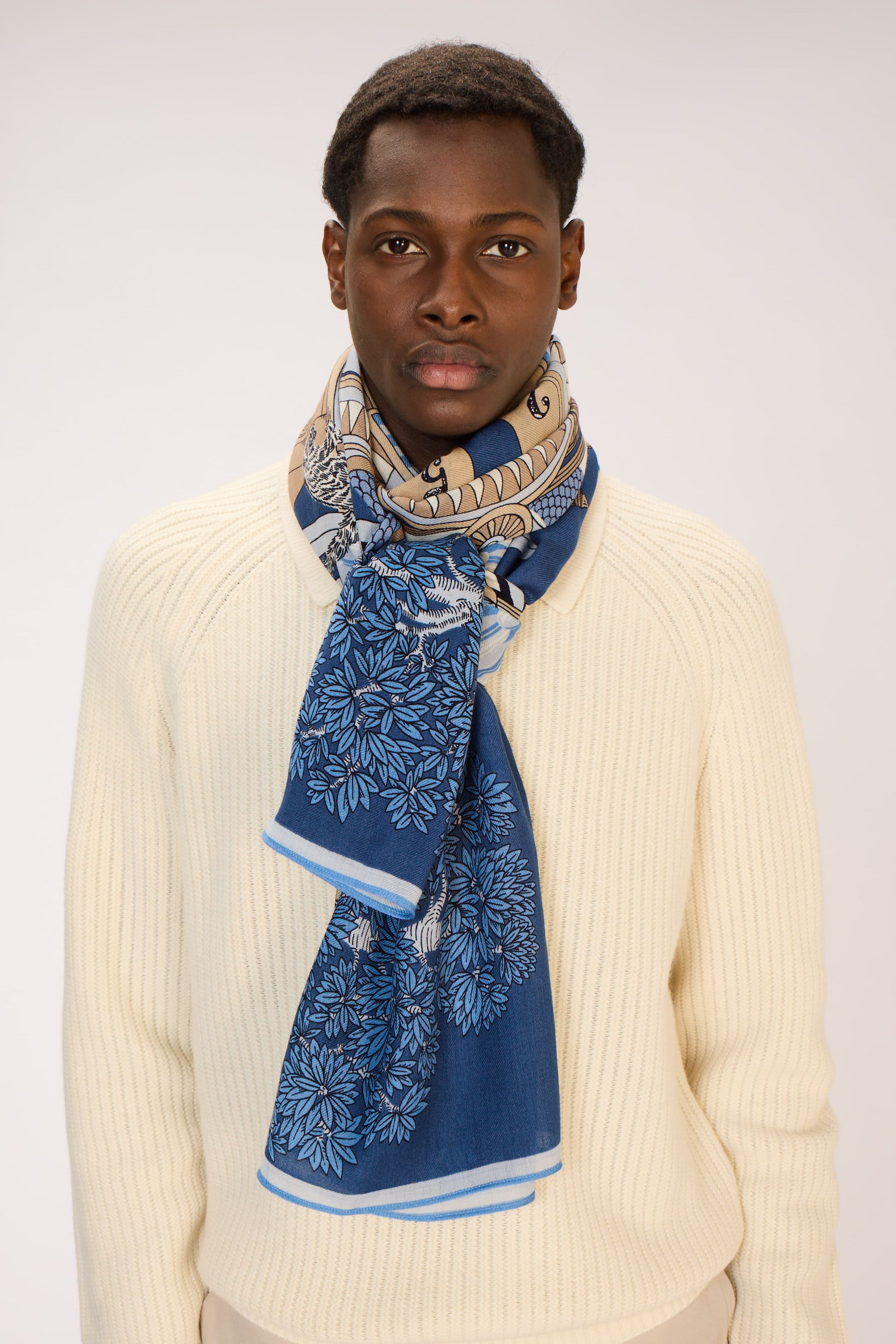 Man wearing a blue patterned scarf and beige sweater on a white background