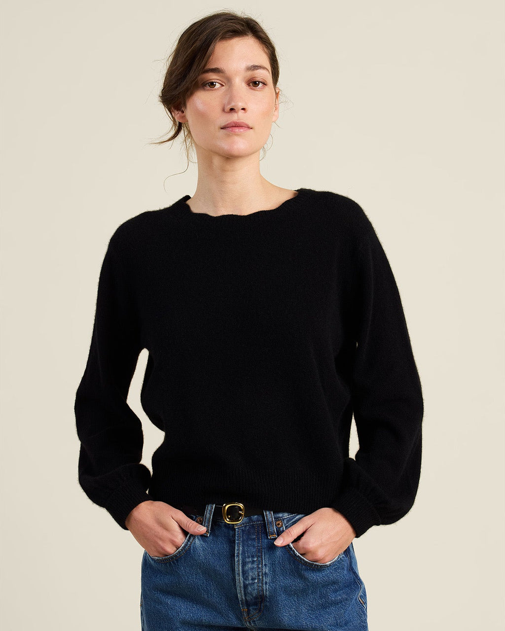 A woman wearing a black long-sleeve knit sweater and blue jeans, standing against a plain light beige studio background.