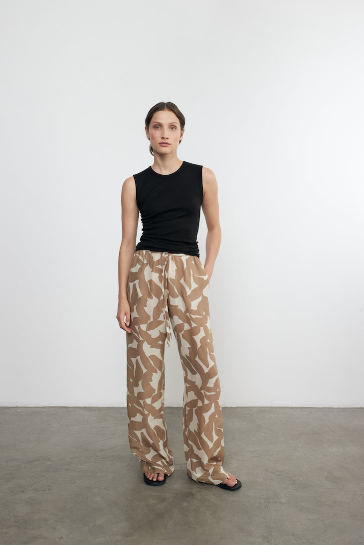Woman wearing a black sleeveless top and patterned pants standing against a white wall.