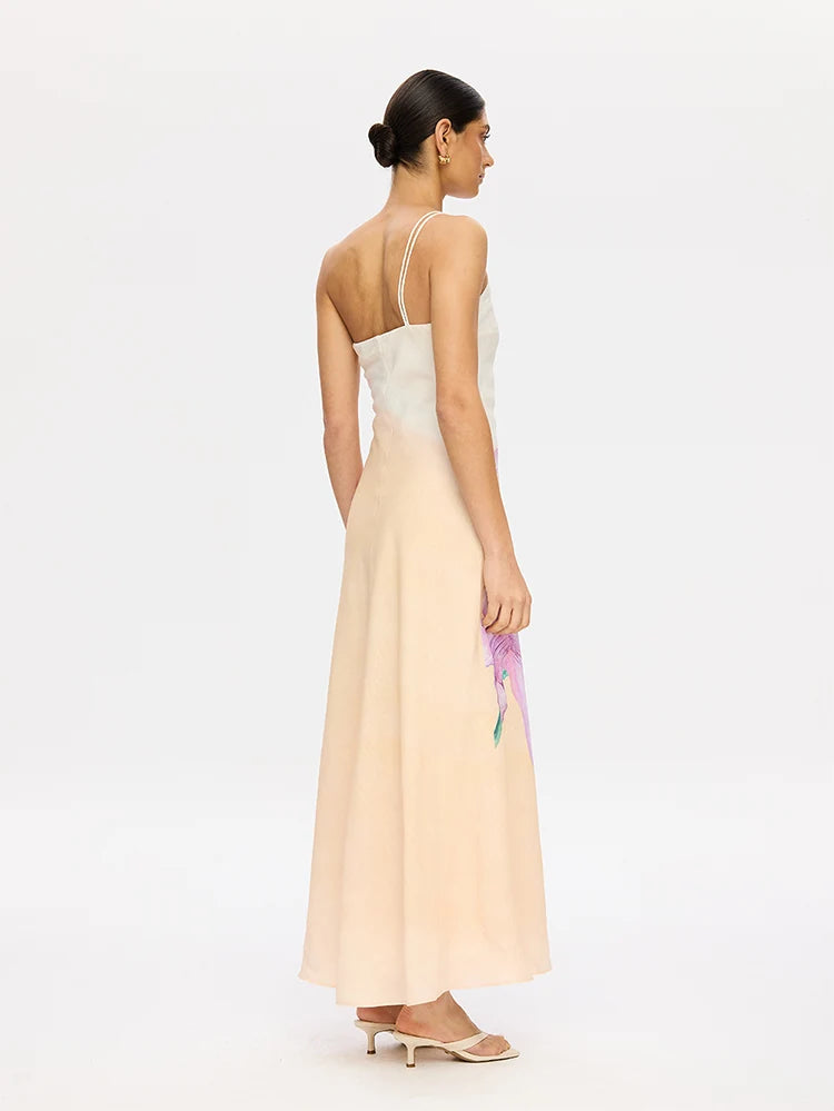 A woman shown from the back wearing a sleeveless maxi dress with thin straps, a fitted bodice, and a flowing pale blush skirt. She is standing against a clean white studio background and holding a small purple flower.