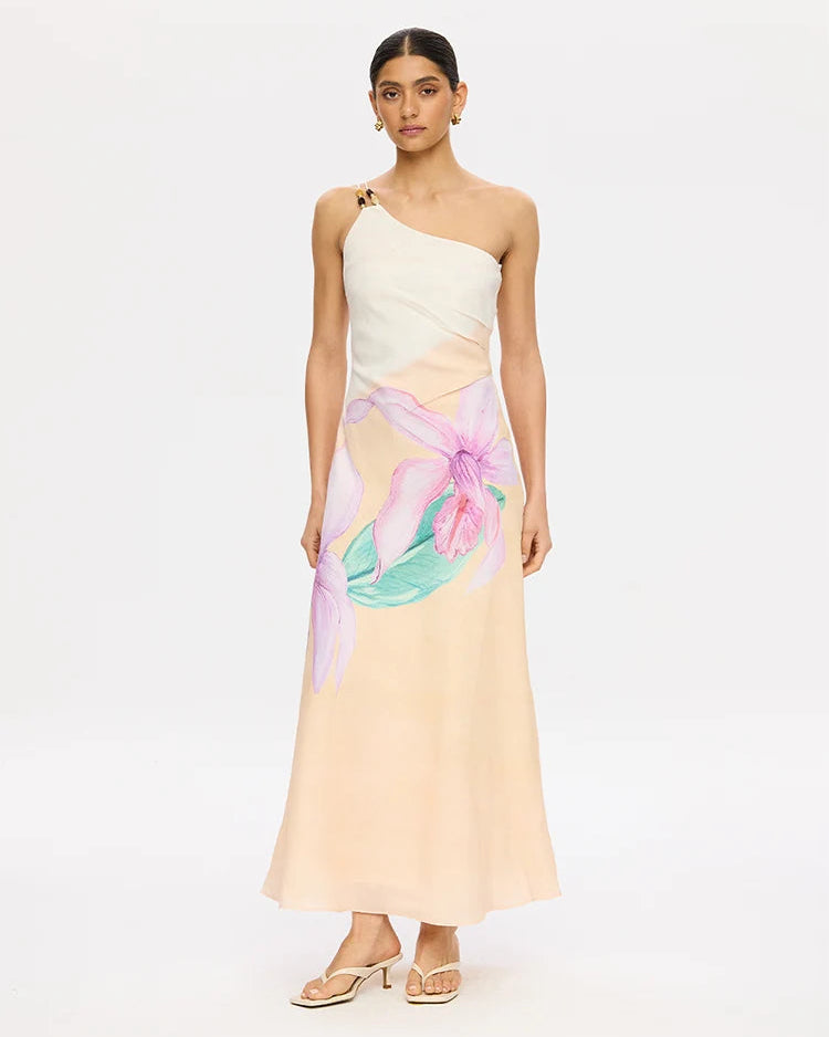 A model wears a one-shoulder maxi dress with a cream upper bodice and a flowing pale peach skirt featuring a large pink and green floral print. She is wearing light beige heeled sandals against a plain white studio background.