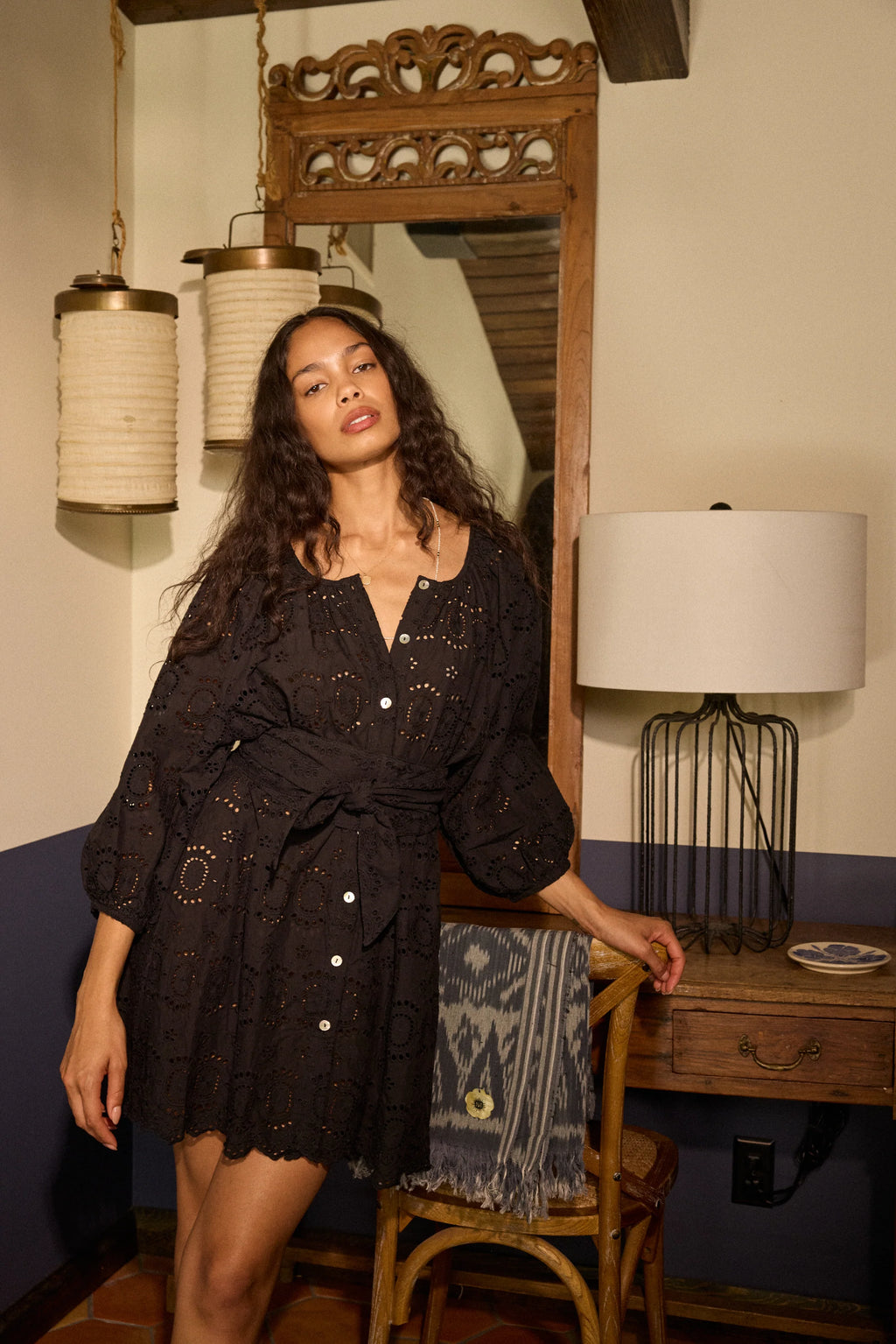 Woman in a black dress standing in a room with a wooden desk and lamp.