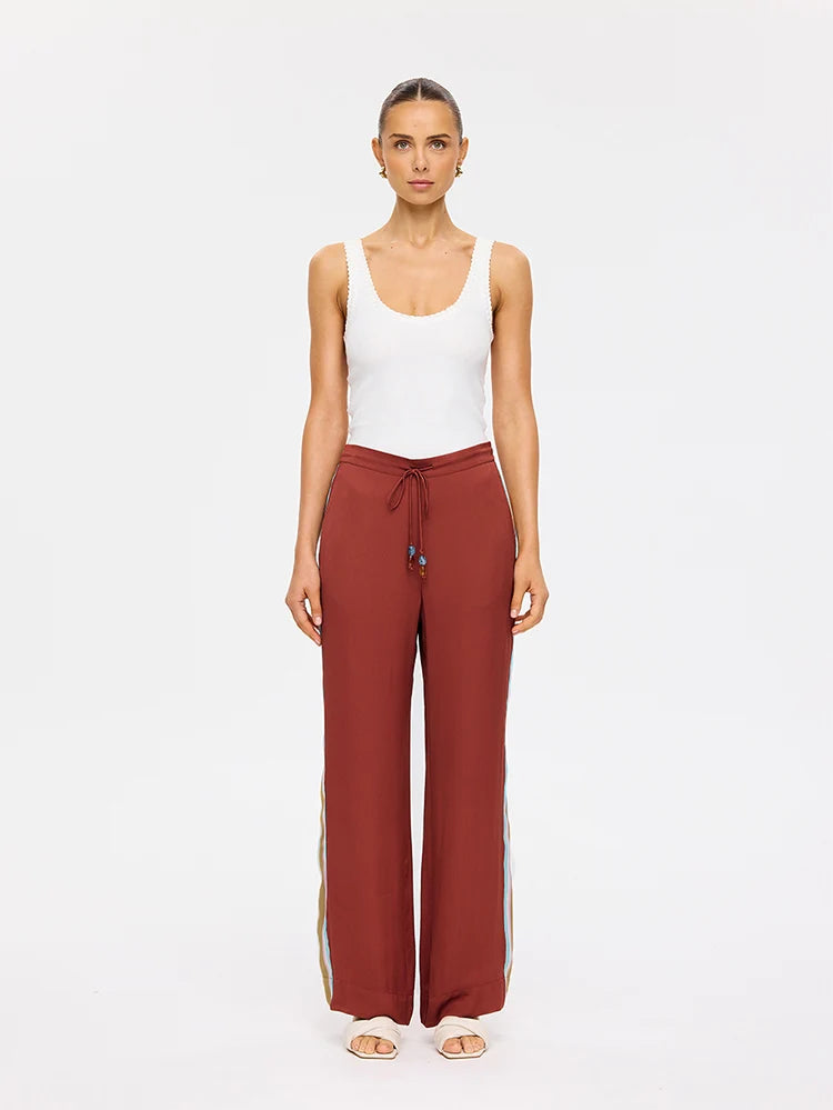 A model stands against a plain white studio background wearing a white sleeveless top and rust-colored wide-leg drawstring pants with thin light-colored side stripes.