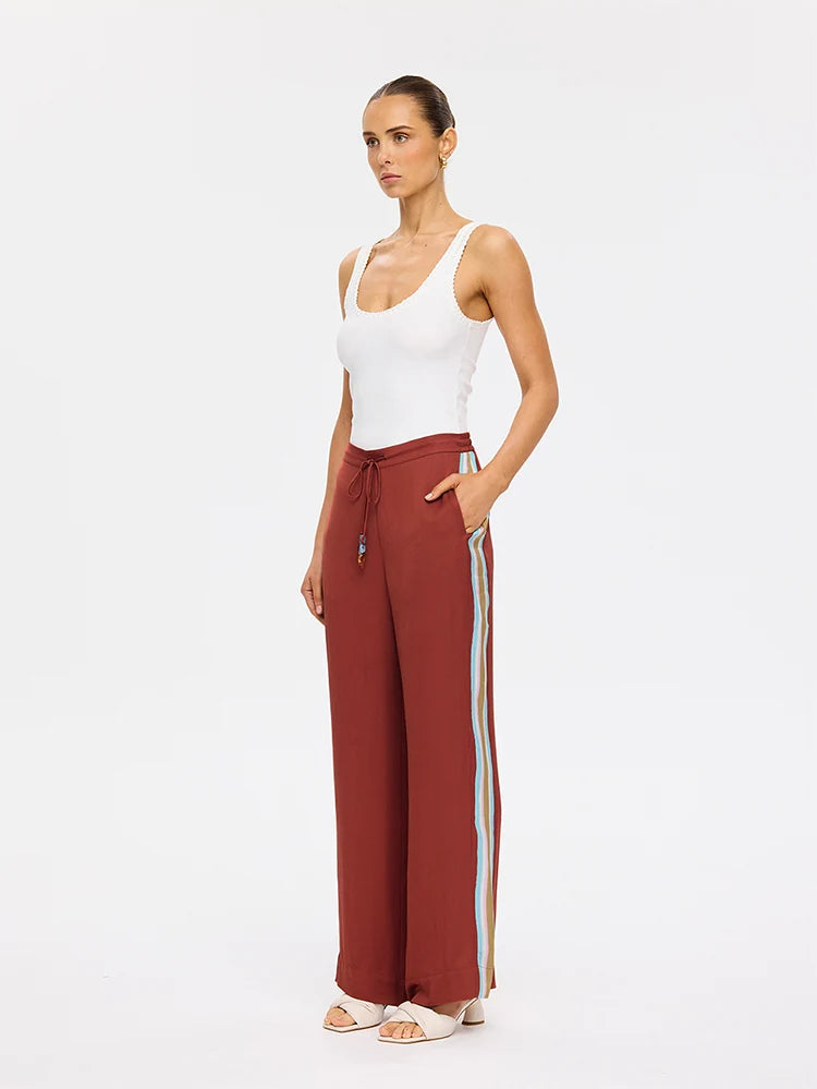 A woman modeling a pair of rust-brown wide-leg pants with light blue and beige side stripes, paired with a white tank top and white sandals, against a plain white studio background.