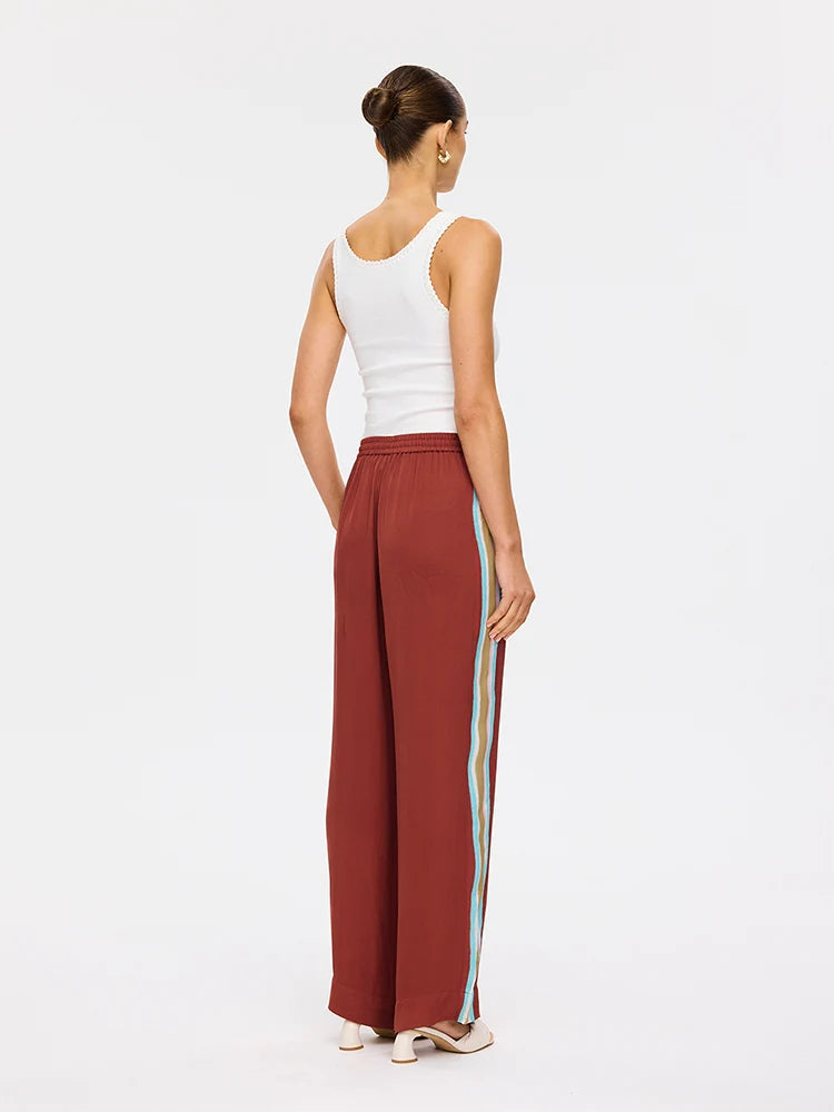 A woman shown from the back wearing wide-leg rust-colored pants with a vertical light blue and yellow stripe down the side, paired with a white tank top and beige heels, against a plain white studio background.