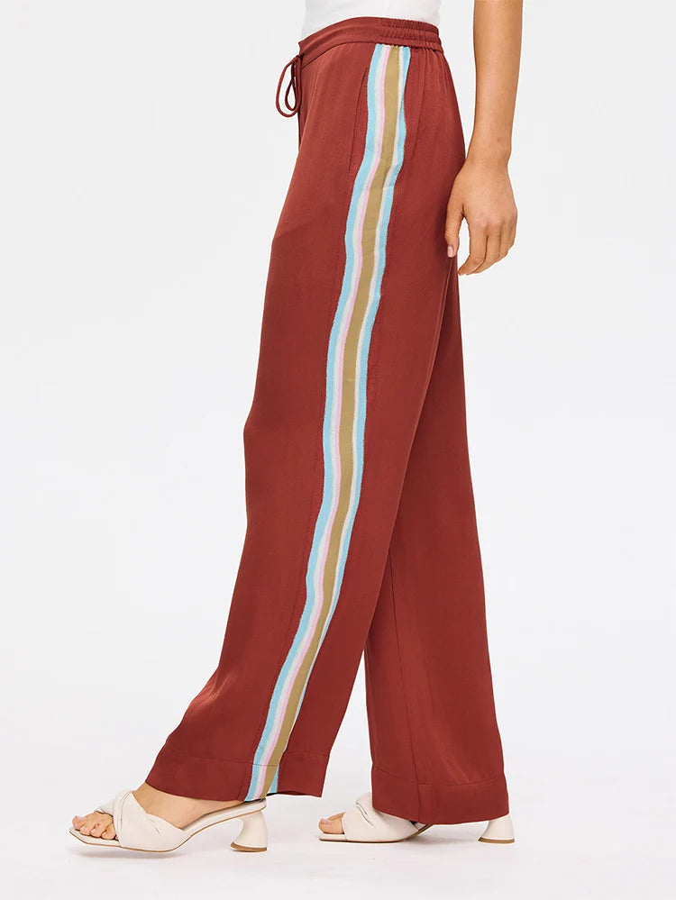 A person wearing rust-colored wide-leg pants with a vertical multicolor stripe down the side, paired with white heeled sandals, shown against a plain white studio background.
