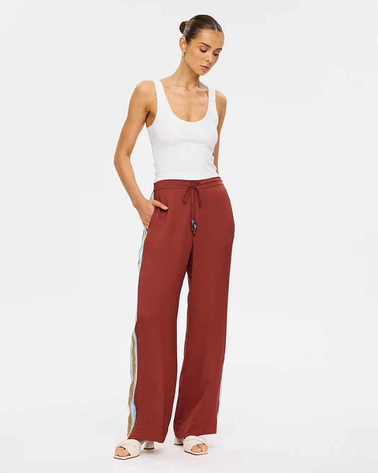 A woman modeling wide-leg rust-colored pants with a side stripe detail, paired with a white tank top and white slide sandals, against a plain white studio background.