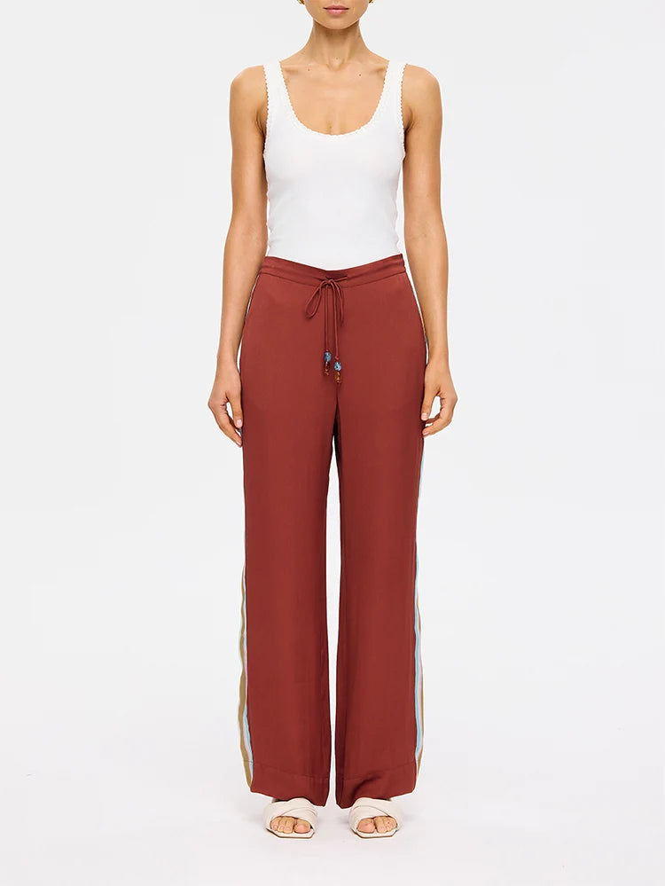 A model wearing rust-colored wide-leg drawstring pants with gold side stripes, paired with a white tank top and white sandals, against a plain white background.