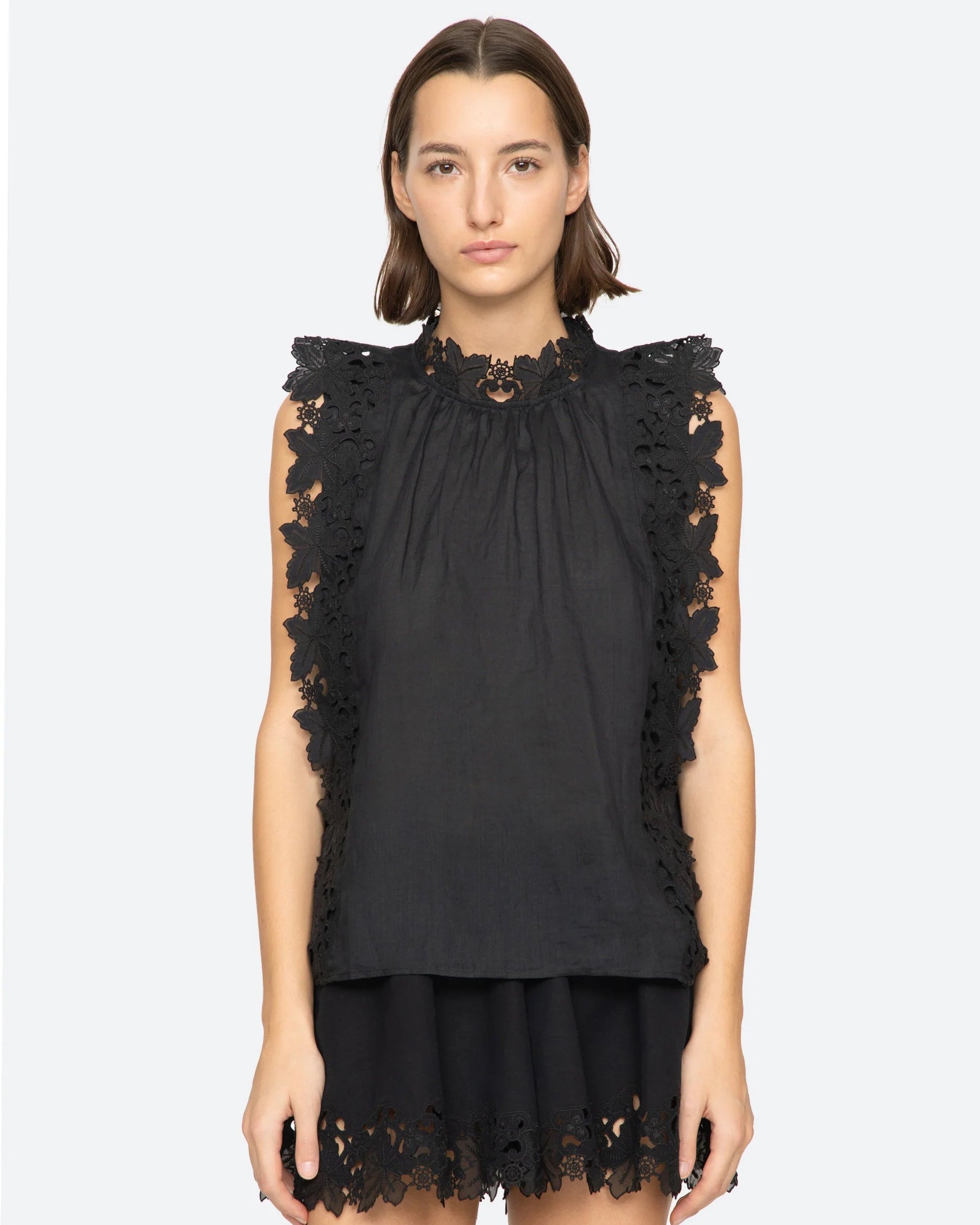 A woman modeling a black sleeveless top with dramatic ruffled lace trim at the shoulders, neckline, and hem, paired with a matching black skirt, against a light gray studio background.