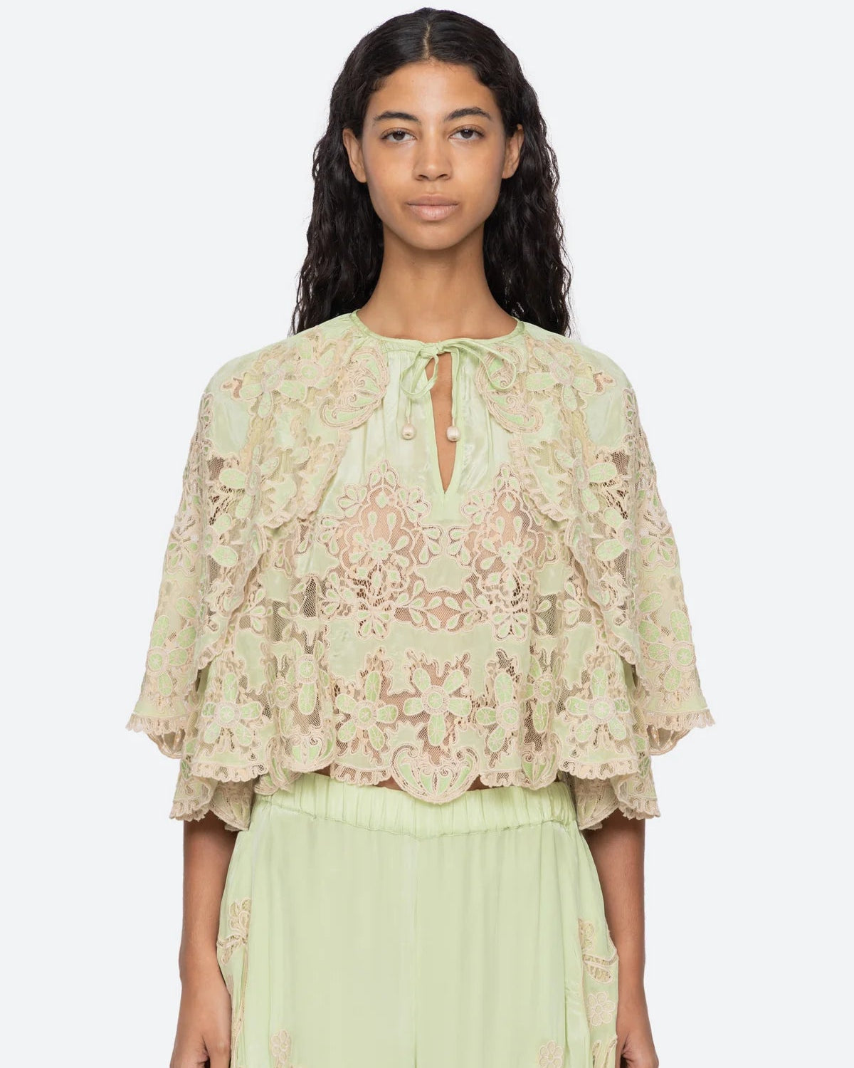 A model wearing a pale sage green lace top with flutter sleeves and a tie neckline, paired with matching sage green pants, against a light gray studio background.
