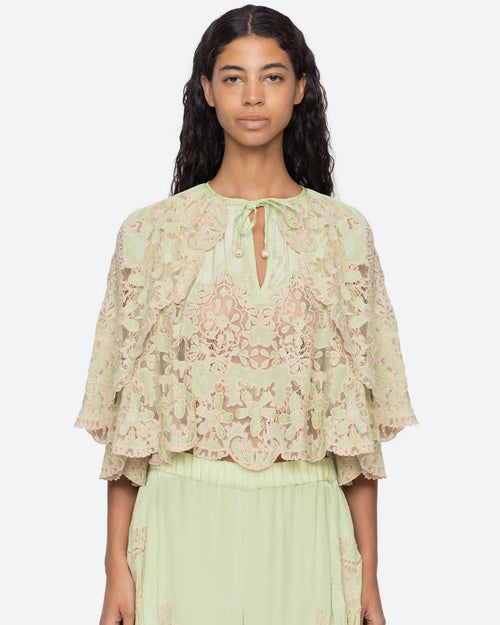 A model wearing a pale sage green lace top with flutter sleeves and a tie neckline, paired with matching sage green pants, against a light gray studio background.