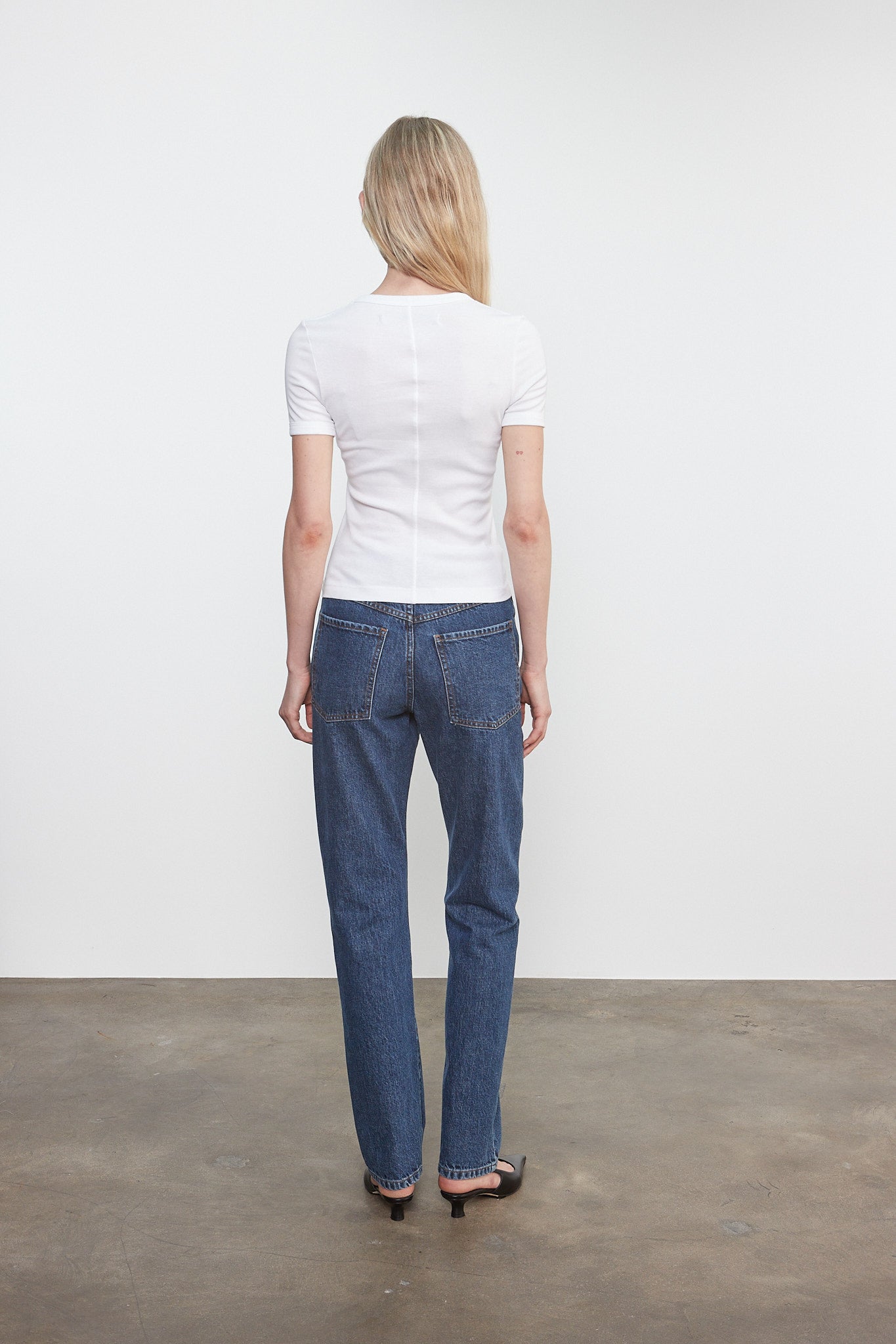 Person wearing a white shirt and blue jeans standing against a plain white wall.