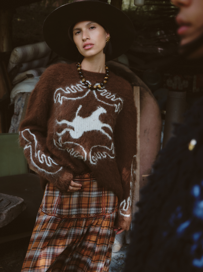 Person wearing a brown sweater with white patterns and plaid pants in a dark setting