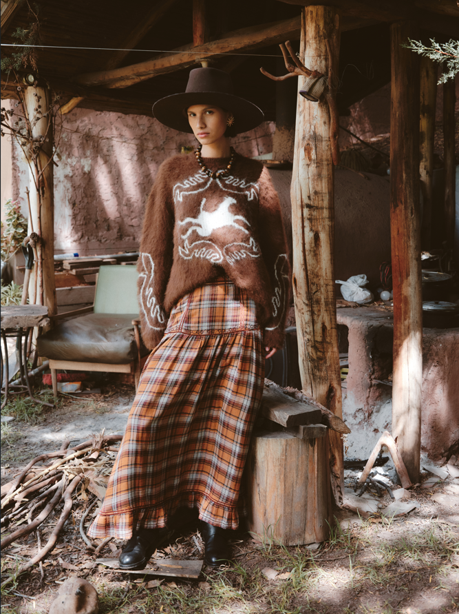 Person wearing a brown sweater with a horse design and plaid pants, standing in front of a rustic wooden cabin.