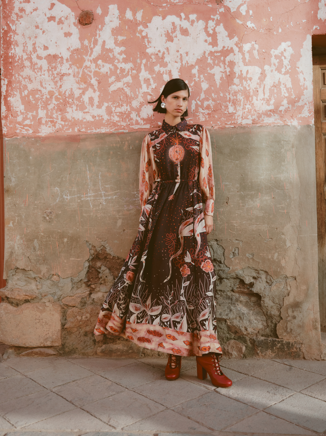 Woman in a patterned dress standing against a weathered wall.