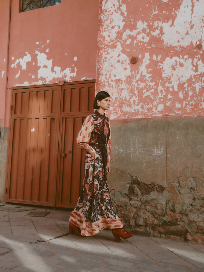 Woman in a floral dress standing against a textured wall with peeling paint.