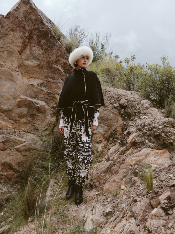 Person wearing a black coat, white fur hat, and patterned pants standing on rocky terrain.