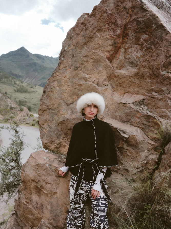 Person in a black outfit with white patterns standing against a rocky background with mountains in the distance.