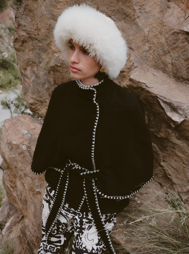 Person wearing a black coat with white trim and a white fur hat against a rocky background